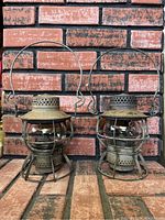 Two vintage Pennsylvania train lanterns made of metal with glass globes and wire handles, placed against a brick wall.
