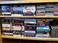 Photo showing multiple VHS tapes stacked on shelves including movies like Titanic, Forrest Gump, Jurassic Park, The Firm, Doctor Zhivago, and boxed sets of Ken Burns Jazz series and Civil War series. CDs stacked above in jewel cases.