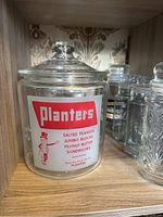 Front view of large Planters peanuts bar glass container with vintage label and fitted lid on wooden shelf.