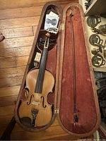 Open hard wooden violin case with reddish-orange interior showing full-size violin inside with bow and some sheet music/instructions.