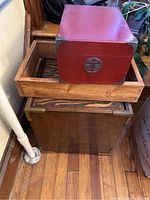 Red wooden box with metal corner braces and circular emblem on front, photographed sitting inside a larger wooden box.