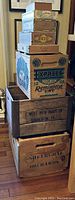 Stacked wooden crates and cigar boxes arranged vertically showing visible brand markings and text from orchards, dairy, and shotgun shell crates.