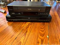 Photo of two Toshiba electronic devices, a VHS player on top and a DVD player below, both black, sitting on a wooden surface.
