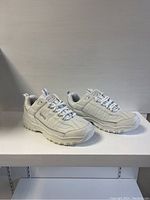 Pair of brand new men's white sneakers displayed side by side on a white surface, showing angled side view with details of laces, sole, and upper material.