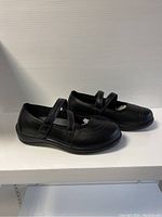 Side view of both black ladies shoes with adjustable straps on shelf.