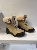 Side view of both boots showing light tan leather and fabric upper with brown rubber soles and toe caps, shearling lining folded over the top and lace-up fronts.