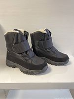 Side angled view of the pair of black unisex winter boots showing the padded upper, strap, sole, and fur lining inside.
