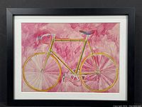 Framed print showing the full yellow bicycle with pink watercolor background in a modern black frame with white matting.