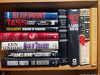 Top-down view of ten vintage spy and detective novels arranged on a wooden surface. Titles include works by Julian Semyonov, Ted Allbeury, Robert Ludlum, and others, mostly hardcover editions with dust jackets.