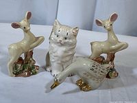 Four porcelain animal figurines including two baby deer, one kitten, and one goose on a white surface.