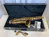 Opened carrying case with saxophone body, case interior, cleaning kit, and instruction booklet visible