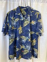 Front view of the blue Tommy Bahama aloha shirt with tropical leaf and flower pattern hanging on a white backdrop.