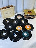 Photo shows approx. 10 loose vinyl 45 RPM records with a visible paper album cover labeled 'Record Album' and a stack of similarly sized records, some in sleeves.