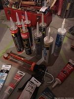 Photo shows assortment of caulking tubes including black, white, red tubes, and an orange caulking gun tool on floor.