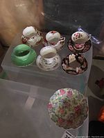 Set of six teacups with saucers, displaying various floral designs and gold trim, on clear plastic surface.
