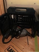 Black & Decker corded drill driver and bit set inside open black plastic carrying case, showing drill with cord, several drill bits, spade bits, and driver bits stored in molded slots.