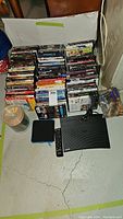 Photo of stacked assorted DVD movies with cases arranged in rows, a spindle of blank or recorded discs, Samsung Blu-ray player unit, and a remote control on concrete floor.