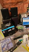 Photo showing the assortment of electronic items including boxed headphones, DVD/VHS player, Philips Blu-ray player box, various speakers, and Logitech accessories.