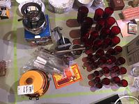 Photo showing a fondue pot on metal stand, boxed fondue kit, metal hand press, stainless steel sifter and an array of red glasses of various sizes arranged on a table.