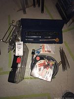 Various tools and accessories including socket set case, rotary tool accessory case, brass faucet valve, files, sharpener pack, and hoses arranged on floor.