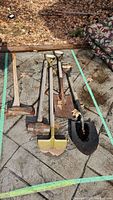 Full view of six outdoor tools laid on stone pavement including one sledgehammer, three shovels, and two axes