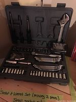 Open black plastic case showing organized socket and wrench set including sockets, wrenches, ratchet wrench, screwdriver handles, and extensions. Tools are metallic with shiny chrome finish.