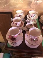 Photo of eleven decorative teacups and matching saucers arranged on a table, showing different floral patterns and gold trim accents.