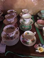 View showing eleven assorted decorative teacups and saucers with floral patterns and different colors, arranged on a tabletop.