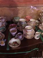 Various teacups, saucers, creamer, and an ashtray arranged on a wooden surface showing assorted designs and colors, with wear visible