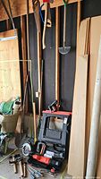 Photo shows different long-handled garden tools including shovels leaning against wall, and smaller hand tools on the floor with power tools nearby.