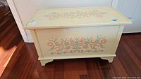 Front view of solid wood toy box showing floral painted design on cream colored lid and bracket feet base