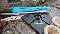 Full view of the disassembled backyard shade umbrella with turquoise canopy fabric resting on the black umbrella base with four connected water-fillable sections on a patio.