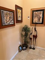 The corner display with the five framed animal prints on the wall, two tall slender wooden animal statues on floor, and artificial plant in a square pot.