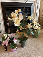 Nine artificial orchid plants grouped on a carpet in front of a fireplace, showing different sizes and colors including white, yellow, and pink flowers in various pots.
