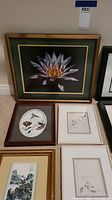 Photo showing a variety of framed pictures arranged on floor: large white flower botanical print with dark green and gold frame, circular floral and bird print in a reddish-brown frame, two minimalist sketches with white mats, traditional Asian landscape in gold frame.