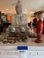 Photo shows an Asian white Buddha statue, two painted decorative eggs in ornately decorated glass boxes, cloisonné egg, brass decorative pieces, and two wooden Japanese dolls.