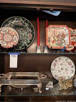 Photo showing four Asian decorative plates in various shapes with floral and dragon designs, two silver-tone candlesticks with red candles, a silver-tone cake plate with lattice design, a serving utensil, and a gold-tone dragon figurine arranged on a shelf.