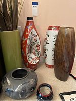 Photo of six Asian vases of different sizes and designs showing red and white floral painted vases, a brown ribbed vase, and round vases on a carpeted floor.
