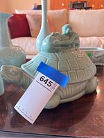 Turtle-shaped celadon jar with figure on top on a wooden table in a living room setting with red and beige couches in background.