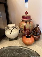 Photo showing five vases of various shapes and designs, including lidded painted vase, white ceramic with rope, small orange round vase, and two brown glazed vases.