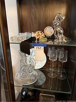 Wide view of mounted glass shelf showing clear crystal pitcher with starburst design, four stemmed wine glasses, clear glass cat figurine, antique salt cellars, and Asian porcelain items in background.