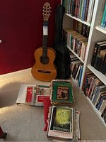 Classical acoustic guitar standing next to black soft case with a bundle of assorted music books on the floor.