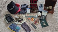 Overview of entire lot on quilted bedspread showing hats, pouches, jewelry pieces, boxes, binoculars, belts, phone sign, adapter, cooling wrap and other items