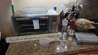 Overview of stainless steel air fryer on countertop with basket on top, knife block, utensil holder, tureen, salt and pepper shakers and additional pan