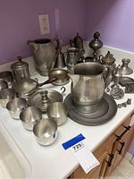 Photo showing a large pewter pitcher, multiple cups, small pan with wooden handle, lidded containers, and decorative pieces grouped on a countertop.