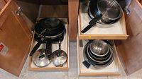 Drawer containing an assortment of pots and pans including frying pans with black handles, saucepans with lids, and metal mixing bowls nested inside each other.