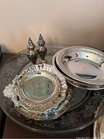 Photo showing pair of ornate salt and pepper shakers, two small serving dishes with handles, and a clear etched glass large charger plate underneath