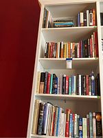 Six shelves of assorted books of various genres and subjects on white bookshelf against red wall