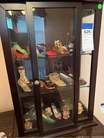 Full view of small dark wood curio cabinet with glass doors, showing collection of miniature shoes on three glass shelves and partial view of glass dragon figurines.