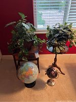 Two wooden tables (one with scalloped edge, one rectangular with medallion inlays), two artificial potted plants with green leaves, and two world globes on carpet in front of window.